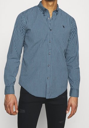 Hombre con camisa de cuadros azul y negra abotonada de manga larga y vaqueros negros rotos sobre un fondo liso.