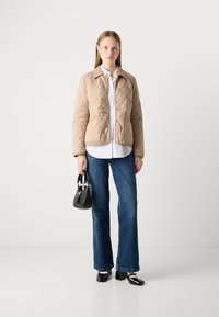 Kvindelig iført beige quiltet jakke, hvid skjorte, blå jeans, sorte lak sko, holder en lille sort håndtaske, står mod en ensfarvet baggrund.