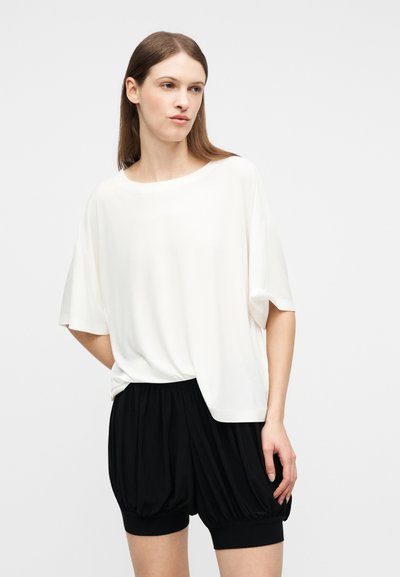Νεαρή γυναίκα με ίσια καστανά μαλλιά φοράει λευκό χαλαρό κοντομάνικο τοπ και μαύρο σορτς με πιέτες, όρθια μπροστά σε απλό φόντο.