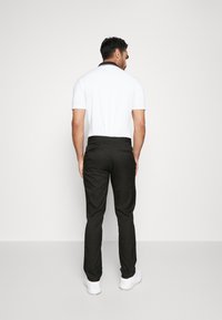 Polo blanc à manches courtes avec col noir, associé à un pantalon noir et des baskets blanches, vu de dos sur un fond uni.