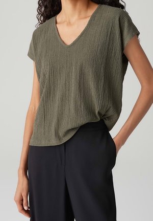 Kobieta w oliwkowozielonej, teksturowanej bluzce z krótkim rękawem i dekoltem w serek, o luźnym kroju, oraz w czarnych spodniach z wysokim stanem, z ręką w kieszeni.