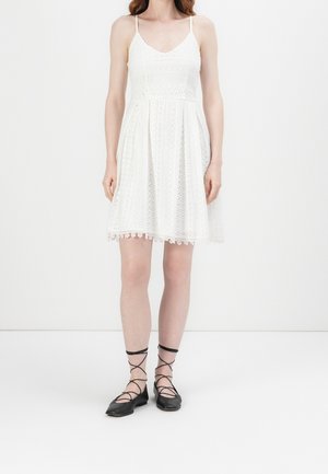 Witte jurk met een aansluitende bodice, kantdetails en een uitlopende rok. Het heeft dunne bandjes en een golvende zoom. Gedragen met zwarte ballerina's.