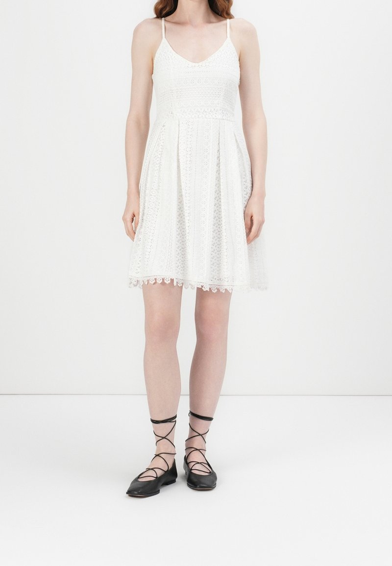 Weißes Kleid mit tailliertem Oberteil, Spitzenverzierungen und einem ausgestellten Rock. Es hat dünne Träger und einen gewellten Saum. Kombination mit schwarzen Ballerinas.