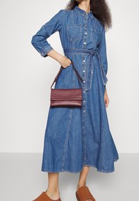 Denim shirt dress featuring a collar, long sleeves, two chest pockets, and a matching belt. Paired with a small burgundy handbag.