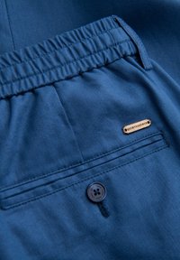 Shorts en tissu bleu avec une taille élastique, une poche arrière et une étiquette indiquant "QUINTESSENCE." Présente une finition lisse et texturée.