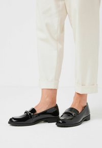 Zwarte gepatineerde instappers met een gladde afwerking, voorzien van een kettingaccent aan de voorkant en een lage, gestructureerde zool. Gedragen met cropped crèmekleurige broeken.