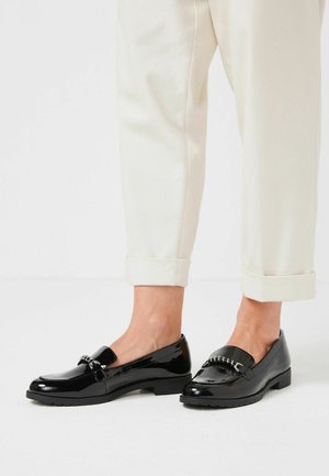 Zwarte gepatineerde instappers met een gladde afwerking, voorzien van een kettingaccent aan de voorkant en een lage, gestructureerde zool. Gedragen met cropped crèmekleurige broeken.