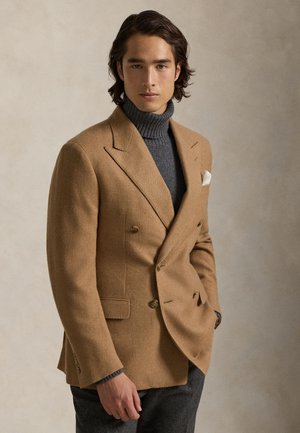 Homme portant un blazer croisé couleur chameau sur un pull à col roulé gris, debout sur un fond beige uni.