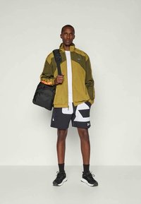 Mann med oliven- og sennepsfarget jakke, svart-hvite rutete shorts, svarte sneakers, bærende en svart skulderveske mot en ensfarget bakgrunn.