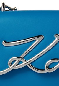 Modrá syntetická kabelka s hladkým povrchem, zdobená výrazným stříbrným logem v kurzívě a metalickým řetízkem.