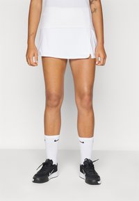 Fehér sport szoknya, harang alakú kivitelben, fekete cipővel és fehér Nike zoknikkal párosítva. Sima anyag; oldalsó hasíték a mozgékonyság érdekében.