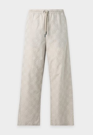 Høj mand iført en beige kortærmet skjorte med knapper og matchende vidde bukser med sorte og hvide sneakers, stående med hænderne i lommerne.