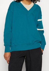 Teal sweatshirt med djup V-ringning, långa ärmar och vita ränder på armarna. Mjuk textur och avslappnad passform.