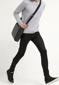 Un pantalon slim noir à la texture lisse, associé à un pull gris clair et des baskets noires. Un sac messenger noir est porté en travers du corps.