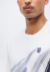 Κοντινό πλάνο ενός ατόμου που φοράει λευκό πουκάμισο K-Swiss με μπλε διαγώνιες ρίγες και στρογγυλή λαιμόκοψη.