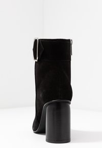 Botín negro de ante hasta el tobillo con un tacón alto y grueso. Cuenta con una correa ancha con una hebilla metálica rectangular. Textura suave y lisa.