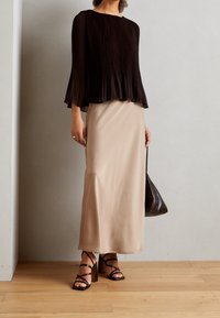 Schwarze plissierte Bluse mit Glockenärmeln kombiniert mit einem beigefarbenen Maxirock aus Satin. Schwarze hohe Sandalen mit Riemchen und eine braune Tasche runden das Outfit ab.
