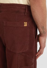 Bordeaux corduroy broek met een gestructureerde afwerking, voorzien van een achterzak en een tan merklabel op de tailleband voor identificatie.