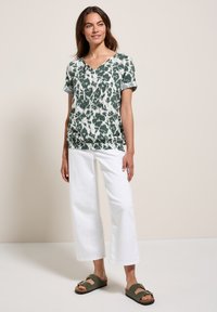 Top corto a maniche corte con motivo verde e bianco e scollatura a V, abbinato a pantaloni bianchi a gamba larga e sandali di pelle verde oliva.