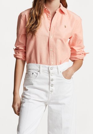 Licht koraalkleurig overhemd met opgerolde mouwen, voorzien van een klein geborduurd logo. Gepaard met hoge witte denim broeken met knoopdetails.