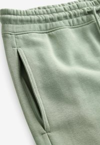 Joggings en coton vert avec une taille élastique et des poches latérales. Le tissu a une texture lisse avec une couleur uniforme.