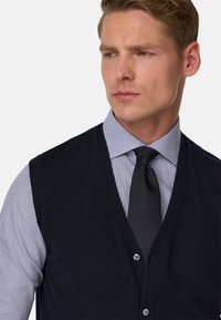 Námořnická pletená vesta s výstřihem do V, nošená přes světle modrou a bílou proužkovanou košili, doplněná tmavým kravatou. Jemné akcenty knoflíků.