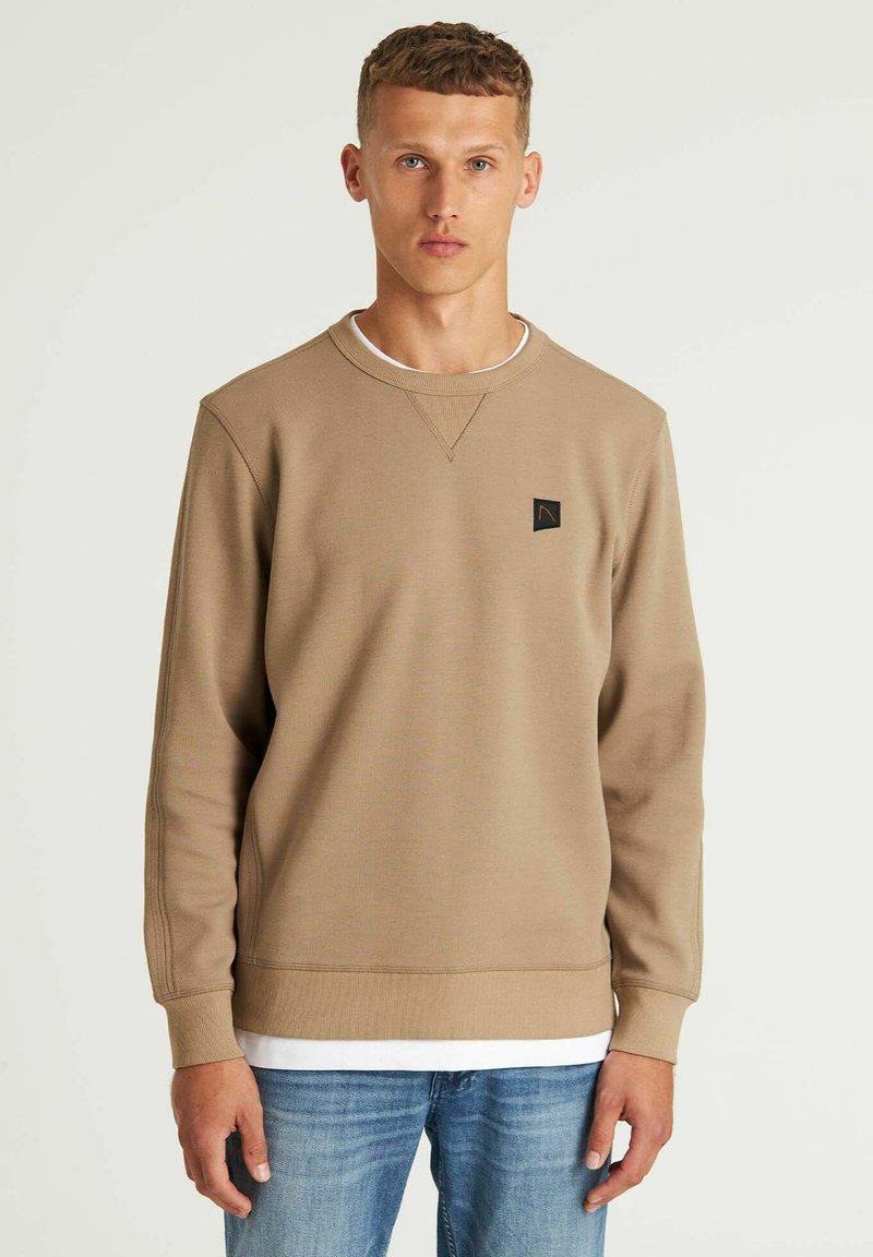 Sudadera marrón de mezcla de algodón, con cuello redondo, puños acanalados y un pequeño parche negro con el logo en el pecho. Llevada sobre una camiseta blanca.