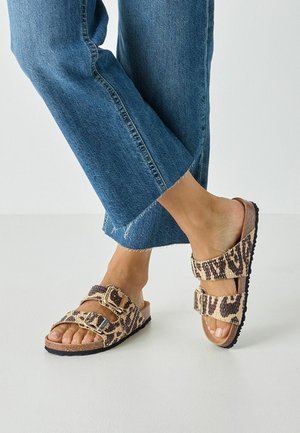 Leopard print slide sandals with two adjustable buckles, cork-like footbed, and textured black rubber sole, paired with cropped denim pants.