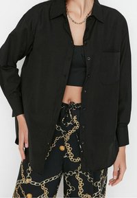 Personne portant une chemise noire ample boutonnée sur un crop top noir, associée à un pantalon noir avec des motifs de chaînes dorées et un collier chaîne dorée.