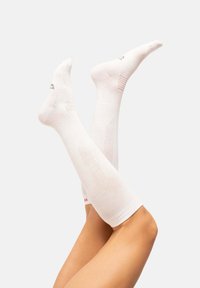 Calcetines blancos hasta la rodilla con superficie texturizada, que presentan un patrón sutil y un pequeño logo rojo en el tobillo. Tejido suave con un acabado liso.