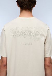 T-shirt couleur crème avec un imprimé graphique des coordonnées géographiques dans le dos. Tissu doux, col rond et manches courtes.