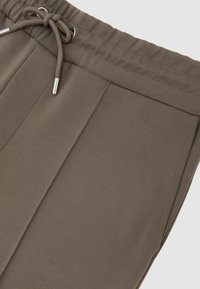 Barna elasztikus derékrésszel ellátott nadrág húzózsinórral, elülső ráncolással és sima textúrával. Látható minták vagy fém részletek nélkül.