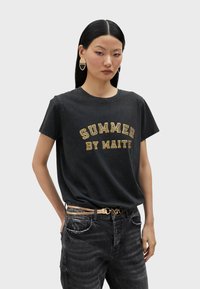 Camiseta negra de manga corta con un acabado desgastado y letras amarillas que dicen "VERANO POR MAITE", combinada con pantalones vaqueros oscuros y un cinturón con detalles dorados.