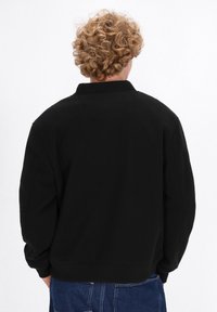 Persona con capelli ricci che indossa una felpa nera e jeans blu, voltata di spalle su uno sfondo bianco semplice.