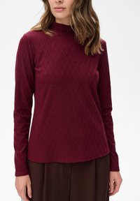 Kobieta ubrana w burgundowy sweter z długim rękawem i wysokim kołnierzem, z delikatnym wzorem w diamenty, oraz ciemnobrązowe spodnie, na jednolitym tle.