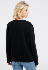 Zwarte sweatshirt met een ronde hals, lange mouwen en geribde manchetten. Heeft een klein logo-label aan de achterkant van de hals. Gestyleerd met blauwe jeans.