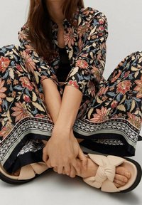 Femme assise en tailleur portant un pantalon noir à motifs floraux et une chemise assortie, avec des sandales beiges ornées de grands nœuds en tissu.