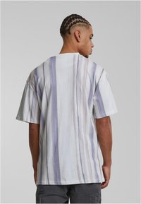 Bő, fehér pólót viselő, copfban hajú férfi, aki háttal áll egy sima szürke háttér előtt, a pólója függőleges szürke és bézs csíkokkal díszített.