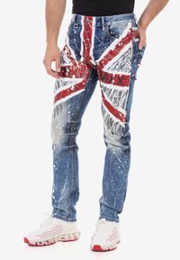 Jean en denim bleu avec un design Union Jack en rouge et blanc, présentant des détails de éclaboussures de peinture et une texture usée. Accentué d'une poche en patch.