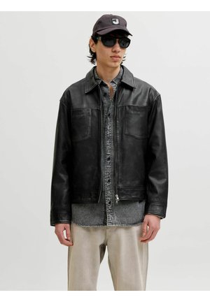 Junger Mann trägt schwarze Lederjacke, graues Hemd mit Knöpfen, hellbeige Hose, schwarze Mütze und dunkle Sonnenbrille vor einfarbigem Hintergrund.