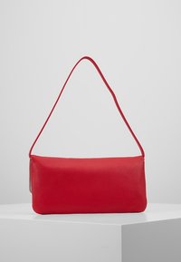 Červená texturovaná kožená taška přes rameno s jedním popruhem, obdélníkového tvaru, vystavená na bílém podstavci proti šedému pozadí.