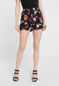 Žena v černých květovaných šortkách se červenými, bílými a zelenými květinami a motýly, spárovanými s černými sandály na vysokém podpatku se zapínáním.