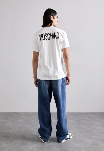 White short-sleeve T-shirt with "MOSCHINO" in black lettering on the back, paired with loose-fit blue denim jeans.