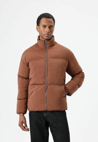 Bruine pufferjas met rits, opstaande kraag, doorgestikte uitvoering en elastische manchetten. Gecombineerd met een donkere spijkerbroek.