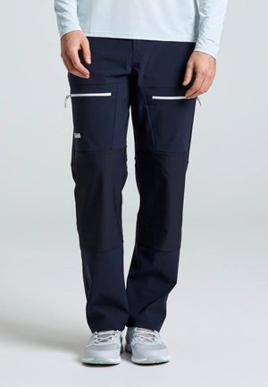 Modelo con pantalones utilitarios azul marino con bolsillos blancos con cremallera en los muslos, una prenda blanca de manga larga y zapatillas grises.