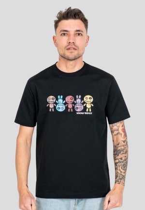 Mężczyzna z tatuażami nosi czarną koszulkę z kolorowymi, naszywanymi postaciami lalek oraz tekstem "WRONG FRIENDS" na jednolitym szarym tle.