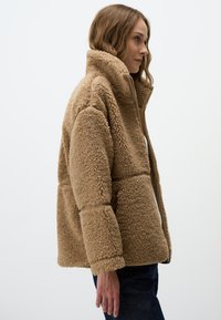 Tan fleece jacket met hoge kraag, ritssluiting en een gestructureerd oppervlak. Heeft een rechte silhouet en lange mouwen.