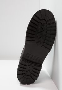 Semelle de chaussure en caoutchouc noir présentant un motif de traction avec des rainures horizontales et verticales. Détail de logo embossé près du talon.