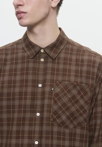 Chemise à carreaux marron à boutons, fabriquée en tissu doux. Présente un col pointu, des boutons blancs et une poche à rabat avec un motif à carreaux diagonal.