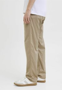 Beiget puuvillahousut, joissa on rento istuvuus ja sivusaumat, yhdistetty valkoisiin lenkkareihin ja valkoiseen paitaan, kuvattu sivulta.